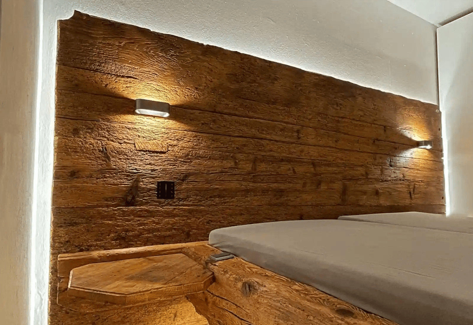 Schlafzimmer mit Altholzwand hinter dem Bett: ein LED-Streifen hinter der oberen Wandkante wirft indirektes warmes Licht an die Decke, zwei kleine Wandleuchten strahlen nach oben und unten.